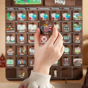 May include: A corkboard calendar with the word "May" at the top. The calendar has a grid of squares, each with a different image representing a daily activity. A hand is holding a square with an image of a movie ticket, popcorn, and 3D glasses. The square says "Movie" at the bottom.