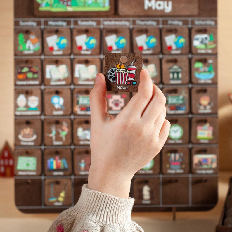May include: A corkboard calendar with the word "May" at the top. The calendar has a grid of squares, each with a different image representing a daily activity. A hand is holding a square with an image of a movie ticket, popcorn, and 3D glasses. The square says "Movie" at the bottom.