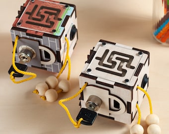 May include: Two wooden puzzle cubes featuring a top-mounted maze, a metal key lock mechanism, and yellow cords with natural wooden beads. The cubes have intricate laser-cut details, with one cube displaying a grey brick pattern and the other a light wood finish.