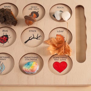May include: A light-colored wooden nature scavenger hunt board with circular cutouts labeled with text and illustrations including BARK, SEED OR NUT, ROCK, BUG, STICK, LEAF, SOMETHING SHINY, SOMETHING COLORFUL, and SOMETHING YOU LOVE, with natural items placed inside.