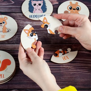 Może przedstawiać: Zestaw sześciu drewnianych puzzli przedstawiających r&oacute;żne zwierzęta, w tym niedźwiedzia, jeża, lisa, wiewi&oacute;rkę i dwa lisy. Każdy element układanki ma kształt innego zwierzęcia i ma nadrukowaną odpowiednią nazwę.