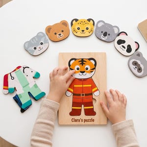 May include: A wooden puzzle board featuring a tiger character dressed as a firefighter, with the text Clara's puzzle printed at the bottom. Additional interchangeable animal heads and clothing pieces are arranged around the central puzzle board.