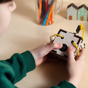 May include: A wooden box with a yellow string threaded through holes. The box has a black switch on the side. A child's hand is pressing the switch.