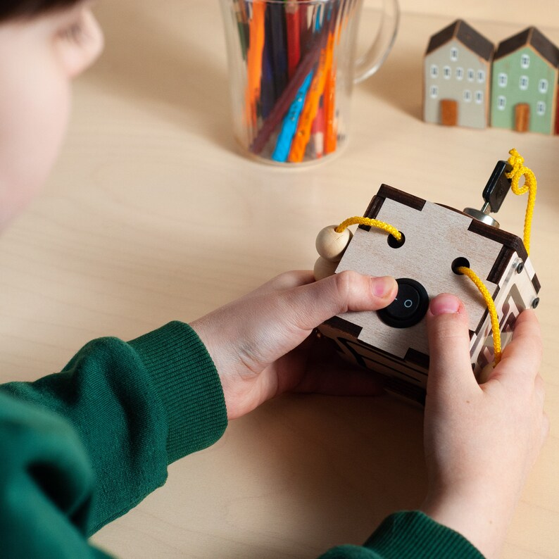 May include: A wooden box with a yellow string threaded through holes. The box has a black switch on the side. A child's hand is pressing the switch.