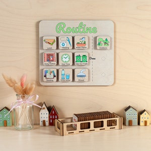 Op de afbeelding: Houten routinebord met kleurrijke pictogrammen voor dagelijkse taken zoals lunch, flossen en afval weggooien. Het woord "Routine" is groen geschreven. Inclusief een houten dienblad met taakkaarten en decoratieve houten huisjes.