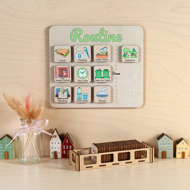 Op de afbeelding: Houten routinebord met kleurrijke pictogrammen voor dagelijkse taken zoals lunch, flossen en afval weggooien. Het woord "Routine" is groen geschreven. Inclusief een houten dienblad met taakkaarten en decoratieve houten huisjes.