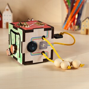 May include: A wooden activity cube with a variety of textures and activities for children. The cube features a light switch, a maze, and a sliding door. It also includes a string with beads that can be threaded through the holes in the cube.