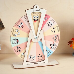 May include: A wooden emotion wheel featuring a rotating pointer and a stationary frame labelled I feel. The wheel is divided into colourful segments, each displaying a cartoon face and an emotion word such as hungry, angry, calm, grateful, relaxed, excited, sick, sleepy, sad, funny, bored, happy, nervous, surprised, and brave.