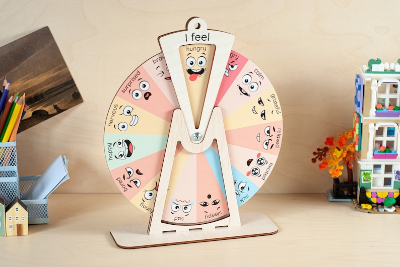 May include: A wooden emotion wheel featuring a rotating pointer and a stationary frame labelled I feel. The wheel is divided into colourful segments, each displaying a cartoon face and an emotion word such as hungry, angry, calm, grateful, relaxed, excited, sick, sleepy, sad, funny, bored, happy, nervous, surprised, and brave.