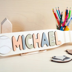 May include: A wooden baseball bat with the name "MICHAEL" spelled out in colorful letters. The bat has a white baseball attached to one end and a stand to hold it upright. There are two small wooden houses and a glass jar of colored pencils in the background.