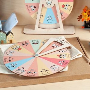 May include: A circular wooden emotion wheel featuring colourful segments with illustrated facial expressions and labels such as happy, bored, and angry. A triangular wooden pointer labelled I feel is attached to the centre, resting on a beige fabric pouch.