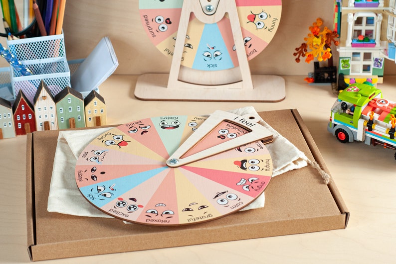 May include: A circular wooden emotion wheel featuring colourful segments with illustrated facial expressions and labels such as happy, bored, and angry. A triangular wooden pointer labelled I feel is attached to the centre, resting on a beige fabric pouch.