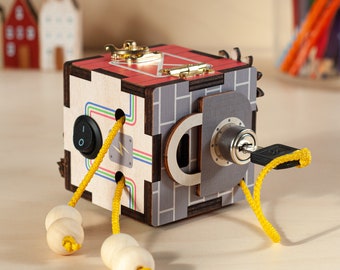 May include: A wooden activity cube with a red, gray, and brown design. The cube has a variety of activities, including a lock and key, a switch, and a maze. The cube is attached to yellow cords with wooden beads.