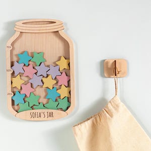 May include: Wooden reward chart shaped like a jar, filled with star-shaped tokens in various colours. The jar has percentage markings. The text "SOFIA'S JAR" is at the base. A wooden hook holds a beige fabric bag.