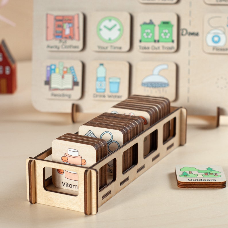 Op de afbeelding: Een houten takenbord met ge&iuml;llustreerde taakkaarten, waaronder "Kleren weg", "Je tijd" en "Vuilnis buiten zetten". Een houten houder bevat extra kaarten met afbeeldingen zoals "Vitaminen" en "Buiten".