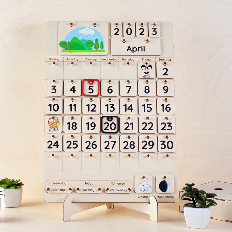 May include: A wooden calendar with a white background and black numbers. The calendar is for April 2023. The calendar has a small picture of a bunny holding a basket of eggs for Easter. The calendar also has a small picture of a cloud with rain drops for weather and a small picture of a crescent moon for moon phase.