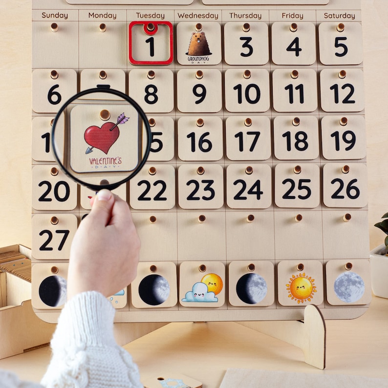 May include: A wooden calendar with a magnifying glass over the date February 14th, which is highlighted with a red heart and the text "Valentine's Day". The calendar has days of the week labeled Sunday through Saturday and numbers 1 through 27.
