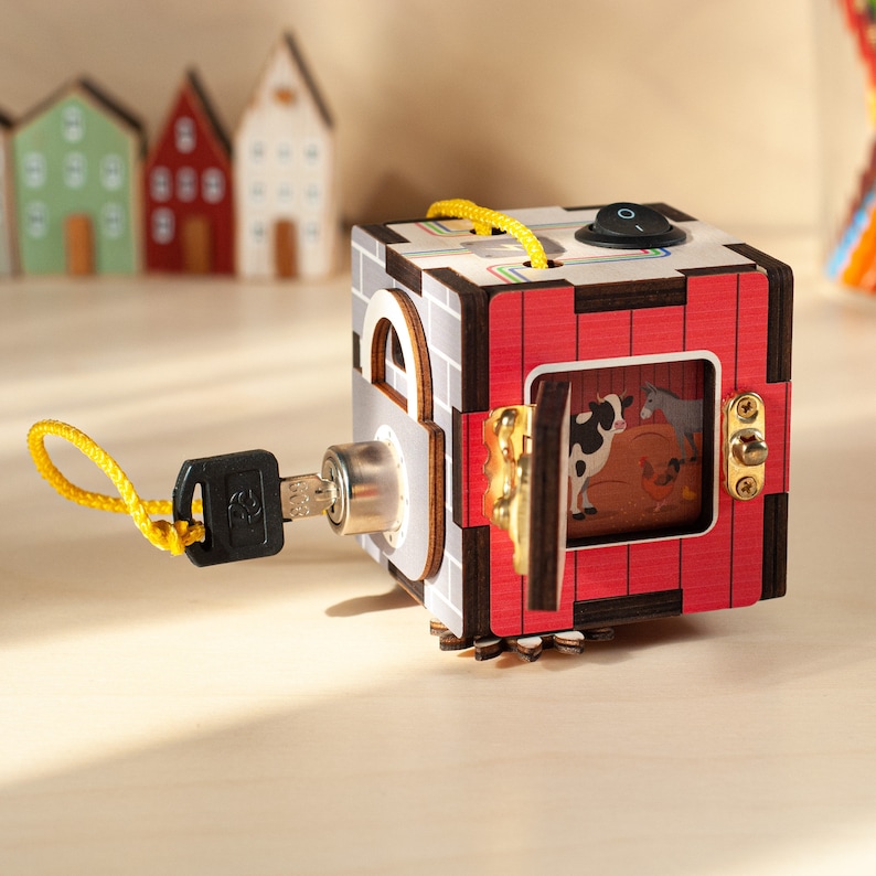 May include: A wooden toy cube with a red door that opens to reveal a farmyard scene with cows and a donkey. The cube has a lock and key, and a yellow string is attached to the key.