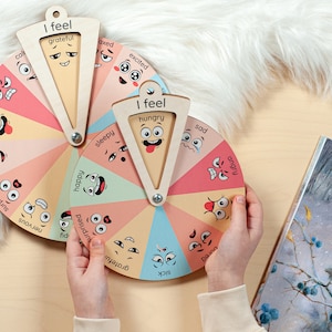 May include: Two circular wooden emotion wheels featuring colourful segments with illustrated facial expressions and labels such as grateful, excited, sad, angry, bored, sick, nervous, surprised, happy, sleepy, brave, and hungry. Each wheel has a rotating pointer.