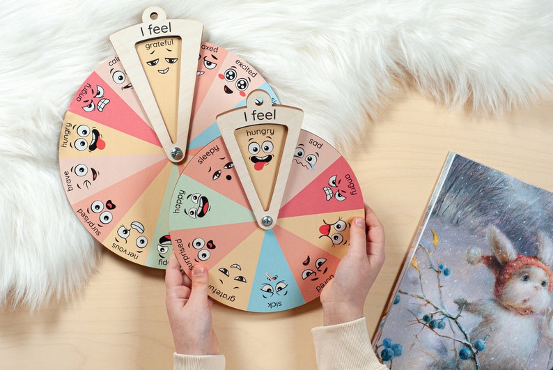 May include: Two circular wooden emotion wheels featuring colourful segments with illustrated facial expressions and labels such as grateful, excited, sad, angry, bored, sick, nervous, surprised, happy, sleepy, brave, and hungry. Each wheel has a rotating pointer.