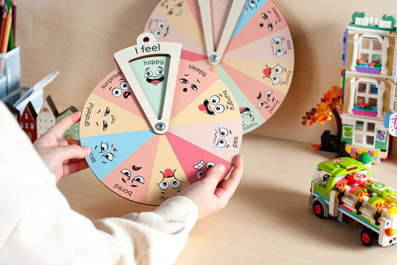 May include: A circular wooden emotion wheel featuring twelve colourful segments, each displaying a unique facial expression and corresponding text labels such as happy, sleepy, hungry, sad, bored, and grateful, with a central pointer labelled I feel.