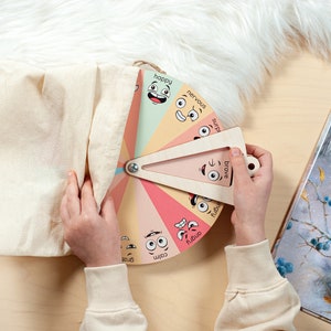 May include: A circular wooden emotion wheel featuring colourful segments with illustrated facial expressions and labels including happy, nervous, surprised, brave, angry, calm, and grateful, paired with a triangular wooden pointer and a beige fabric bag.