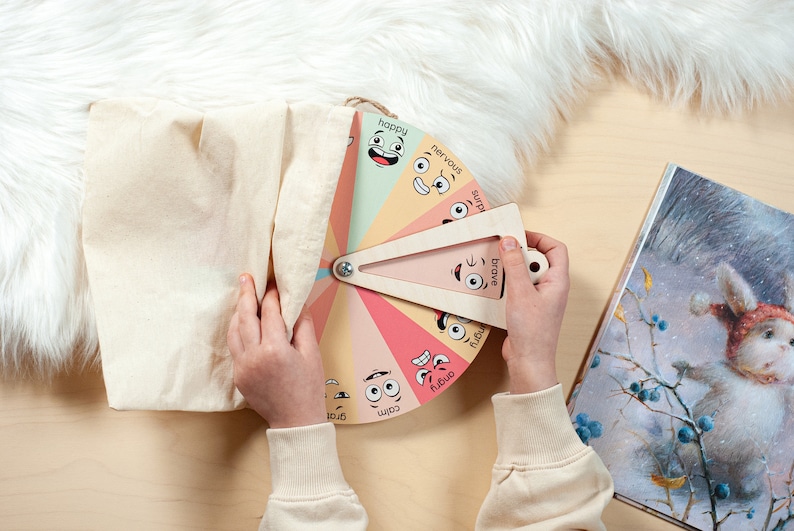 May include: A circular wooden emotion wheel featuring colourful segments with illustrated facial expressions and labels including happy, nervous, surprised, brave, angry, calm, and grateful, paired with a triangular wooden pointer and a beige fabric bag.