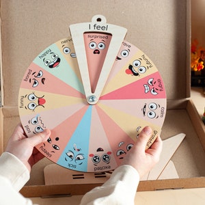 May include: A circular wooden emotion wheel featuring twelve colourful segments, each displaying a unique cartoon face and an emotion label such as happy, bored, funny, sad, sleepy, sick, excited, grateful, calm, angry, hungry, and surprised.