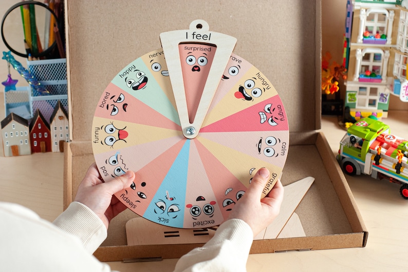 May include: A circular wooden emotion wheel featuring twelve colourful segments, each displaying a unique cartoon face and an emotion label such as happy, bored, funny, sad, sleepy, sick, excited, grateful, calm, angry, hungry, and surprised.