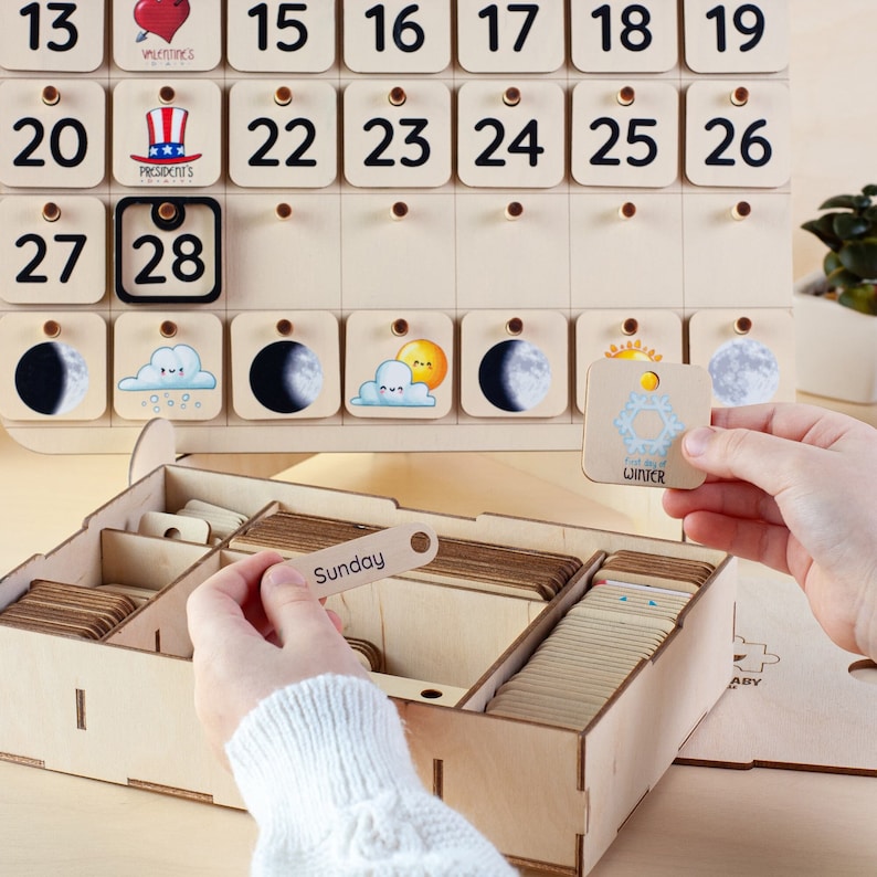 May include: Wooden calendar with removable tiles for days, months, and weather. The calendar includes a box with additional tiles for days of the week, holidays, and weather icons. The tiles feature illustrations of hearts, stars, a hat, a moon, a sun, and a snowflake.
