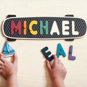 May include: A wooden skateboard-shaped puzzle with the name "MICHAEL" spelled out in colorful letters. The letters are missing from the puzzle and are being placed by a child's hands. A small wooden sailboat is also in the image.