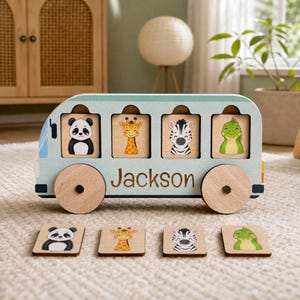 May include: A light blue wooden bus toy featuring the name Jackson printed in brown. The bus has four square cutouts containing illustrations of a panda, giraffe, zebra, and dinosaur. Four matching wooden tiles are placed in front of the bus.