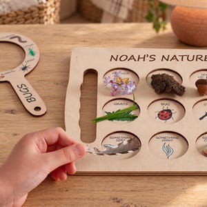 May include: A light-colored wooden nature scavenger hunt board with the text NOAH'S NATURE, featuring circular cutouts for items like a flower, bark, acorn, grass, and a ladybug illustration, alongside a matching wooden magnifying glass.