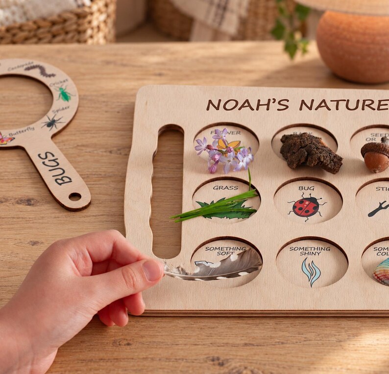 May include: A light-colored wooden nature scavenger hunt board with the text NOAH'S NATURE, featuring circular cutouts for items like a flower, bark, acorn, grass, and a ladybug illustration, alongside a matching wooden magnifying glass.