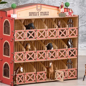 May include: A wooden toy horse stable with multiple stalls, painted red and natural wood. The sign on top reads "Sophia's Stable". The stable is filled with toy horses and figurines. A toy horse stands in front.