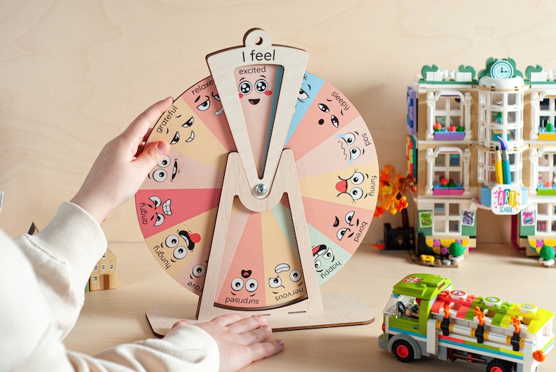 May include: A circular wooden emotion wheel featuring pastel-coloured segments, each displaying a unique facial expression and corresponding label such as excited, sleepy, sad, funny, bored, happy, nervous, surprised, brave, hungry, angry, grateful, and relaxed.