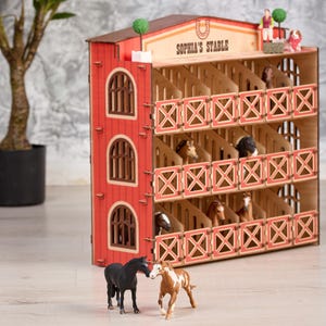 May include: A wooden toy stable with red walls and multiple stalls, labelled "Sophia's Stable." The stable features miniature horse figurines and decorative elements. Two toy horses stand in front of the stable.