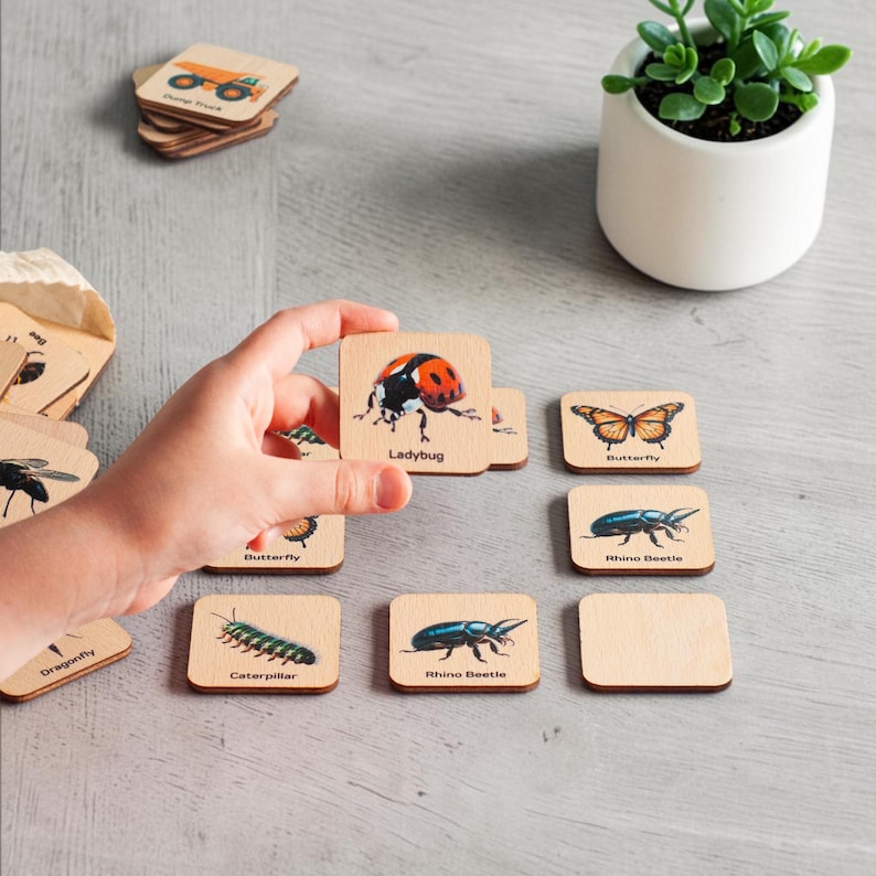 May include: A set of square wooden tiles featuring colorful illustrations of insects and animals with their names printed below each image. The tiles display a ladybug, butterfly, rhino beetle, caterpillar, and dragonfly on a light wood surface.