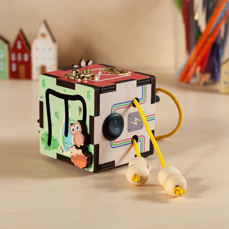 May include: Wooden activity cube with a variety of textures and activities for children. The cube features a green and brown side with a maze, a red and white side with a sliding door, and a side with a light switch and a lightning bolt. The cube also has two yellow cords with wooden beads.