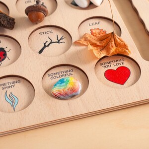 May include: A light-colored wooden nature scavenger hunt board with circular cutouts. The board features printed text and illustrations including 'STICK', 'LEAF', 'SOMETHING COLORFUL', 'SOMETHING YOU LOVE', and 'SOMETHING SHINY', with a seashell and leaf placed on it.