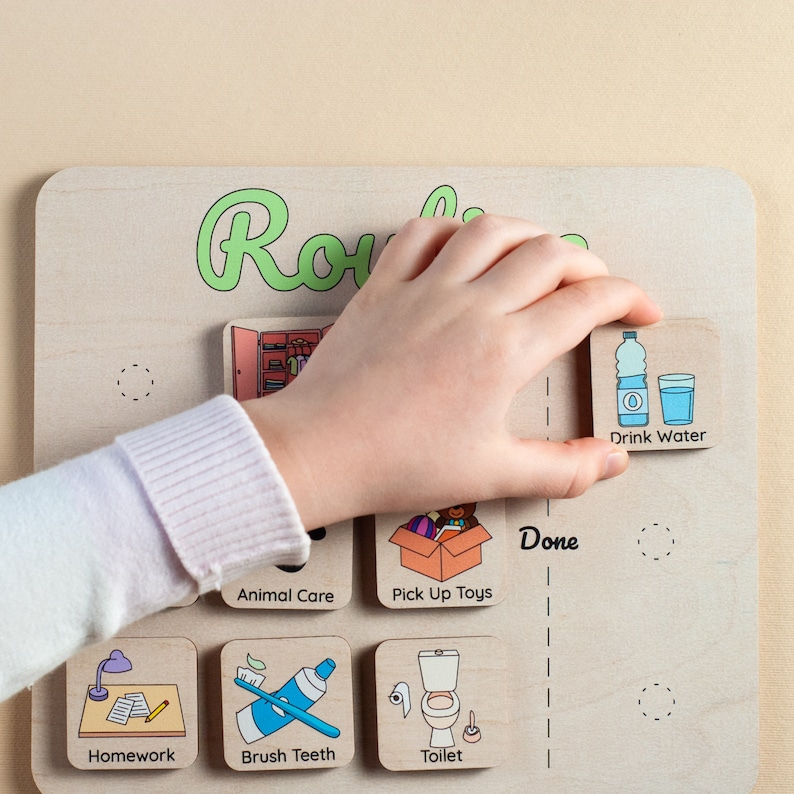 Op de afbeelding: Houten routinebord met ge&iuml;llustreerde activiteit tegels. Een hand plaatst een "Drink Water" tegel. Andere tegels tonen "Huiswerk", "Tanden poetsen", "Toilet", "Dierverzorging" en "Speelgoed opruimen". Het woord "Routine" is groen geschreven.