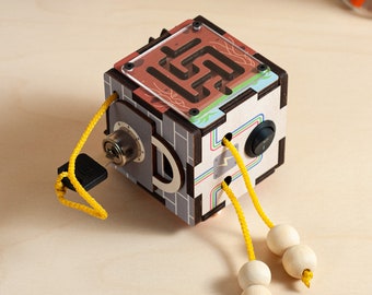 May include: A wooden cube toy with a maze, a lock, and a switch. The cube has a colorful design and is attached to two yellow cords with wooden beads.