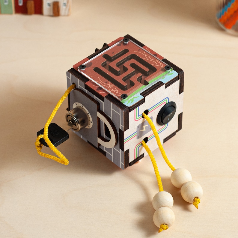 May include: A wooden cube toy with a maze, a lock, and a switch. The cube has a colourful design and is attached to two yellow cords with wooden beads.