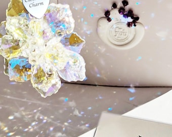 May include: A clear iridescent crystal flower ornament with a silver chain and a heart-shaped charm that says "Customize Charm". The ornament is packaged in a white box with a wax seal that says "For You". The background is a light gray with a sparkly effect.