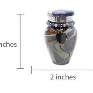May include: A small, decorative urn with a swirling pattern in shades of blue, brown, and gray. It has a dark blue lid and a beaded accent around the neck. A black heart-shaped charm hangs from the neck. The urn is 3 inches tall and 2 inches wide.