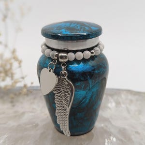 May include: A small, blue and black urn with a decorative lid and a silver charm. The charm features a heart and a wing. A beaded bracelet encircles the urn. The urn is designed to hold a small amount of cremated remains.