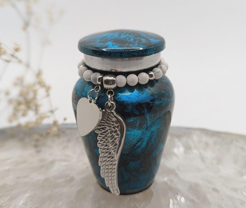 May include: A small, blue and black urn with a decorative lid and a silver charm. The charm features a heart and a wing. A beaded bracelet encircles the urn. The urn is designed to hold a small amount of cremated remains.
