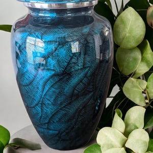 May include: A blue and black cremation urn with a glossy finish. The urn has a lid and a silver band around the top. The surface is decorated with a feather-like pattern. The urn is sitting on a gray stone.