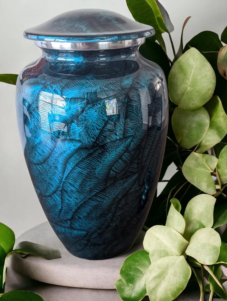 May include: A blue and black cremation urn with a glossy finish. The urn has a lid and a silver band around the top. The surface is decorated with a feather-like pattern. The urn is sitting on a gray stone.