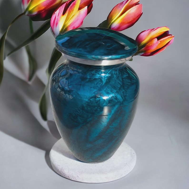 May include: A teal-colored urn with a silver rim and lid, resting on a white stone base. The urn has a glossy finish with a marbled effect. Colorful tulips with red, yellow, and pink petals are arranged near the urn.